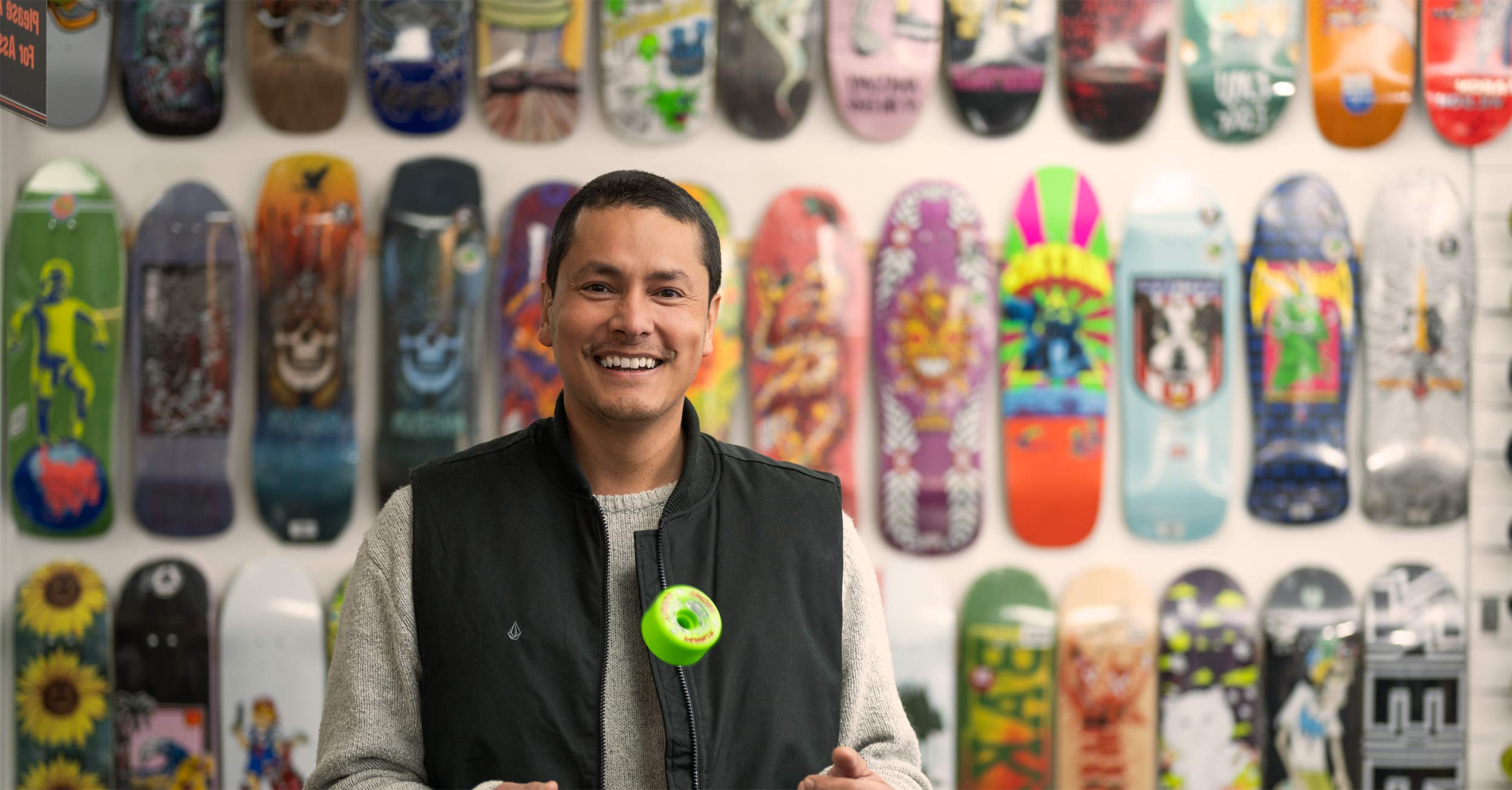 A man smiles at the camera while standing in front of a shop wall covered with skateboards.